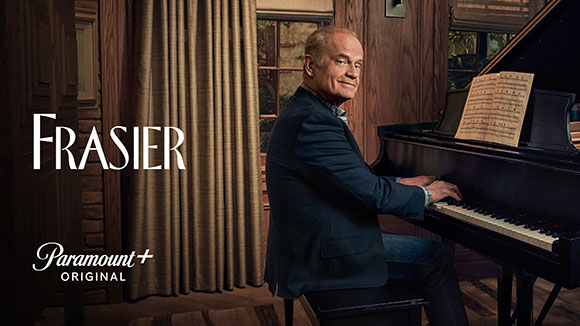 A man in a suit sits at a black grand piano in a room with curtains and a window.