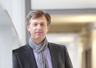 A man wearing a dark blazer, striped shirt, and grey scarf stands in a bright hallway.