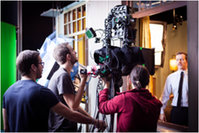 Film crew operating a large camera rig in front of a set featuring a man in a white shirt and tie.