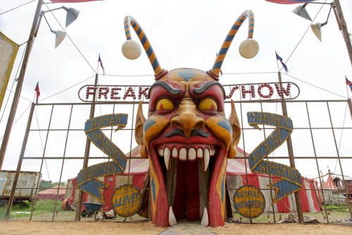 A large, colorful demon mask with long antennae and sharp teeth marks the entrance to a freak show.
