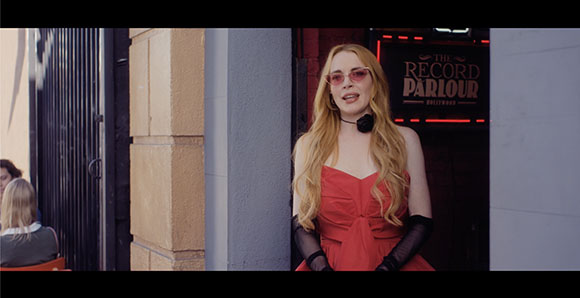 A woman with long blonde hair and pink sunglasses wearing a red dress stands before a club entrance.