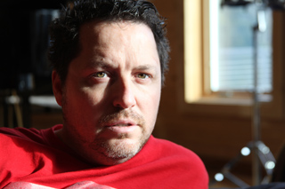 Close-up of a man with dark hair and stubble wearing a red shirt, looking toward the camera.