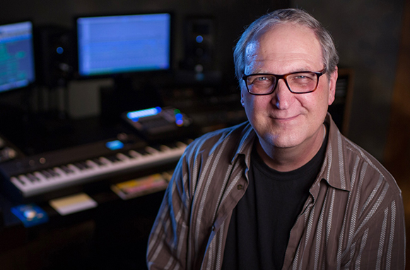 A man with glasses and a striped shirt smiling in a music studio with keyboards and monitors.