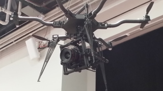 A large drone with multiple rotors and a professional camera mounted underneath is suspended by wires from an indoor ceiling.