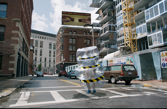 A person in a large, clear inflatable suit stands in a city street near parked cars and construction.