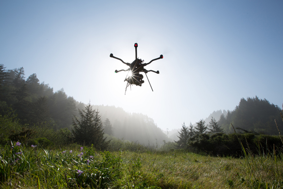 Silhouette of a multi-rotor drone flying against a bright sun over a foggy forest landscape.