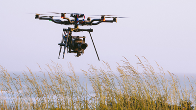 A large black drone equipped with a professional cinema camera flies over a field of tall, golden grass.