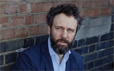 A man with dark curly hair and a beard wearing a blue blazer and light blue shirt.