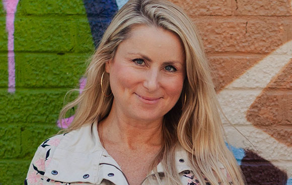 A smiling blonde woman wearing a patterned jacket in front of a colorful brick wall.