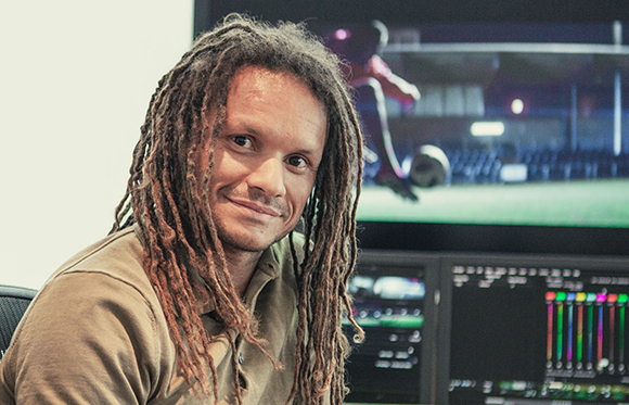 A man with long dreadlocks smiles while sitting in front of video editing monitors.