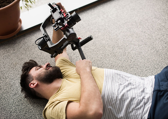 A man lies on a gray carpeted floor holding a professional camera gimbal up toward his face.
