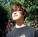 A man with dark hair and glasses looks upward against a background of green trees and foliage.