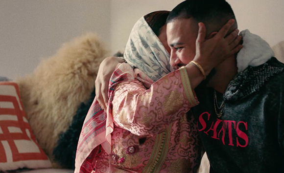 A woman wearing a light blue headscarf and pink patterned tunic hugs a man in a dark sweatshirt.