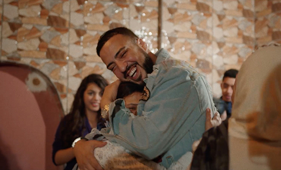 A man with a beard and denim jacket smiles broadly while embracing another person.