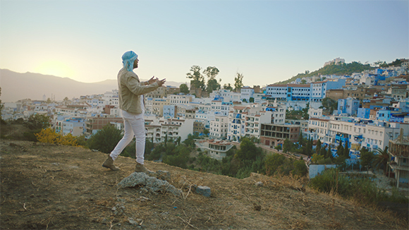 A man in a light jacket and headwrap stands on a hill overlooking a town with blue buildings.
