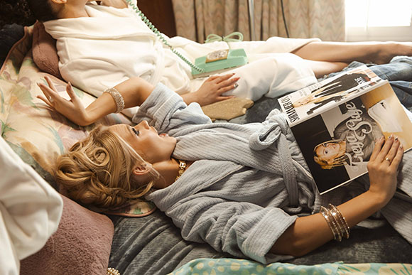 Two women in robes lounge on a bed; one holds a magazine while the other talks on a vintage telephone.
