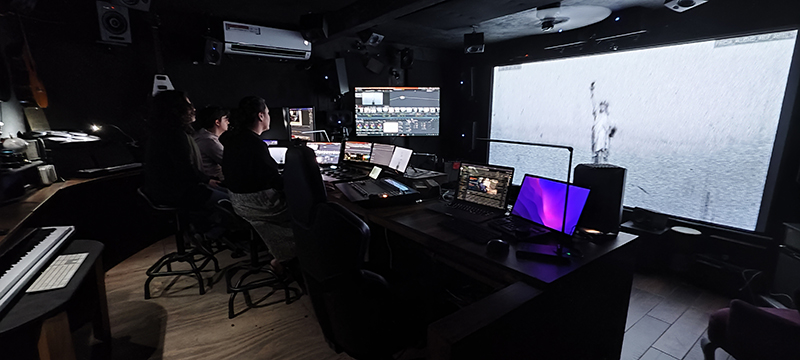 Three people sit in a dark studio facing multiple computer monitors and a large projection screen.