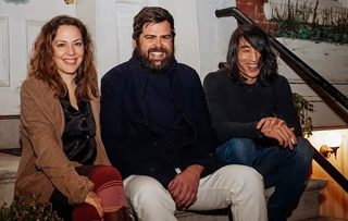 A woman in a brown jacket, a bearded man in a dark coat, and a man with long hair sit together on steps.