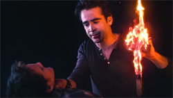 A man holds a bright, vertical flame near the face of a person lying down in a dark setting.