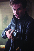 A man in a dark jacket holds a handgun with a focused expression.