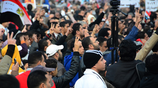 A dense crowd of people gathered outdoors, with several individuals raising their hands in the air.