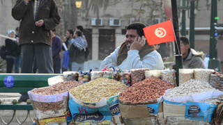 A man sits behind a street vendor cart overflowing with bags of assorted nuts and snacks.