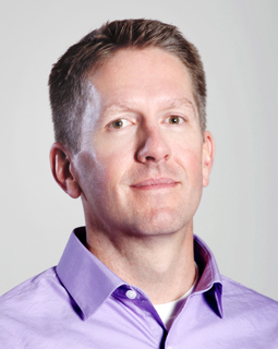 A man with short brown hair wearing a light purple button-down shirt against a plain light background.