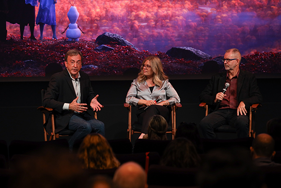 Three people sit on chairs in front of a screen displaying characters from Frozen 2, including Elsa, Anna, and Olaf.