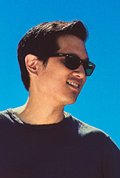 A man with dark hair and sunglasses smiling against a bright blue sky.