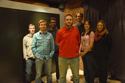 Seven diverse people standing in a group in a room with brick walls and dark curtains.