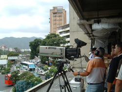 A large video camera on a tripod points toward a city street with buses and buildings under a cloudy sky.