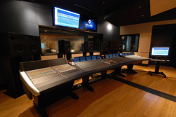 A large audio mixing console sits in a dark studio with monitors and large speakers.
