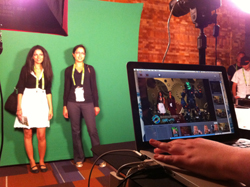 Two women stand before a green screen, with a laptop in the foreground showing a digital composite of people and characters.