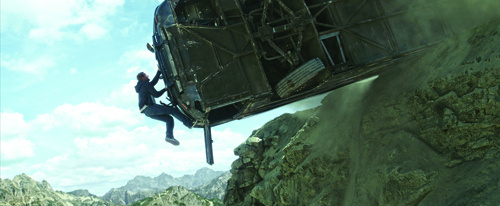 A man hangs from the bottom of a dark, overturned vehicle on a steep, rocky mountainside.