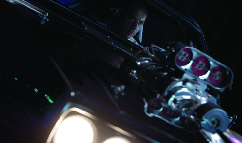 A person sits in a dark vehicle behind a large, chrome engine part with three circular red accents.