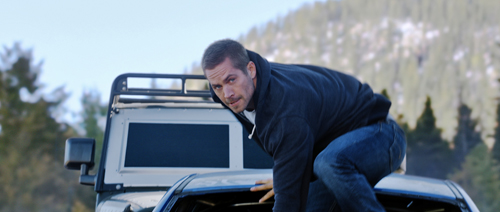 A man in a dark hoodie and jeans leans over the hood of a light-colored vehicle outdoors.