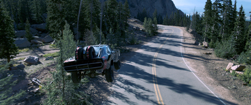 A dark car travels along a winding two-lane road through a mountainous, forested landscape.