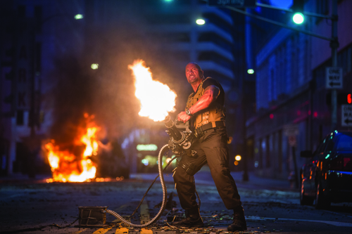 A man in tactical gear fires a large weapon that shoots a massive burst of fire on a dark city street.