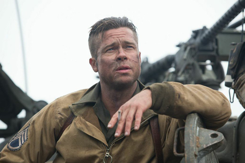 A man in a tan military uniform with dirt on his face leans against heavy machinery.