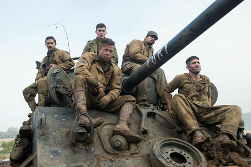 Several soldiers in tan uniforms sit on a mud-covered tank under a cloudy sky.
