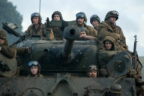 Several soldiers in military gear and helmets ride atop and around a large, muddy tank.
