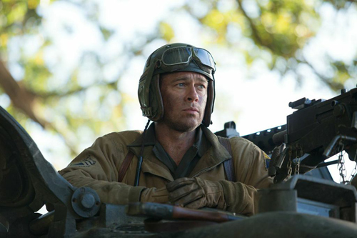 A man wearing a flight helmet and goggles sits in a military vehicle looking forward.