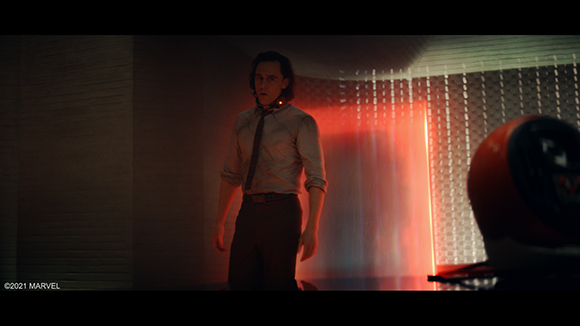 A man in a light shirt and dark trousers stands in a dark room illuminated by a bright red light.