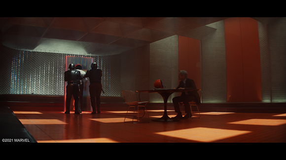 Three armored guards stand by a doorway as a man sits at a table in a dimly lit, modern room.