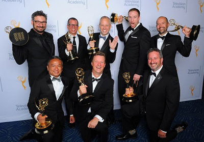 Seven men in black tuxedos pose together holding gold Emmy awards against a white Academy backdrop.