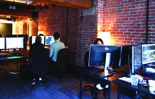 People sitting at desks with multiple computer monitors in a room with red brick walls.