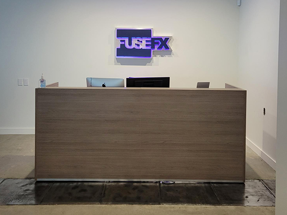 A wooden reception desk in front of a white wall featuring a glowing purple FUSEFX sign.