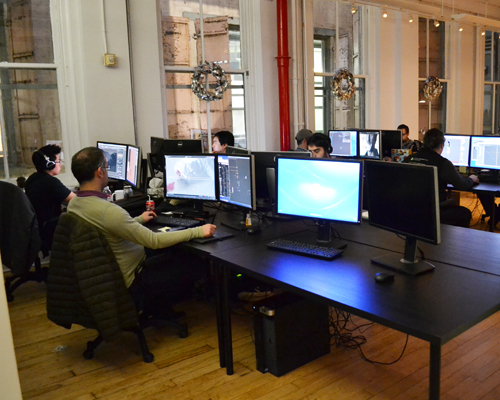 Several people sit at long black desks with multiple computer monitors in a bright, high-ceilinged office.
