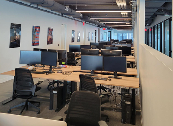 Rows of wooden desks with multiple computer monitors and black ergonomic chairs in a modern office.
