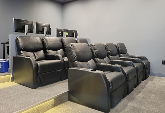 Two rows of black leather reclining theater seats facing monitors on a raised platform.